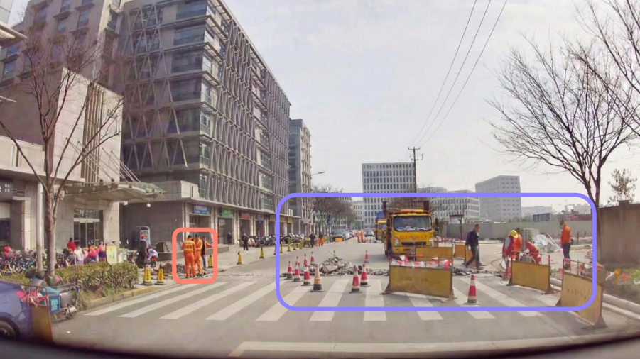 Dashcam frame of an urban road works zone with cones, barriers, a truck and road workers in orange, the workers and the work zone outlined